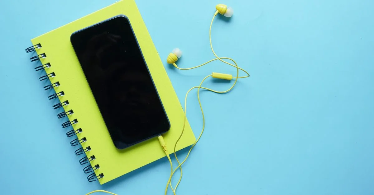 Composition colorée d'un smartphone, d'un cahier et d'écouteurs sur un fond bleu. Parfait pour des thèmes technologiques et de style de vie.