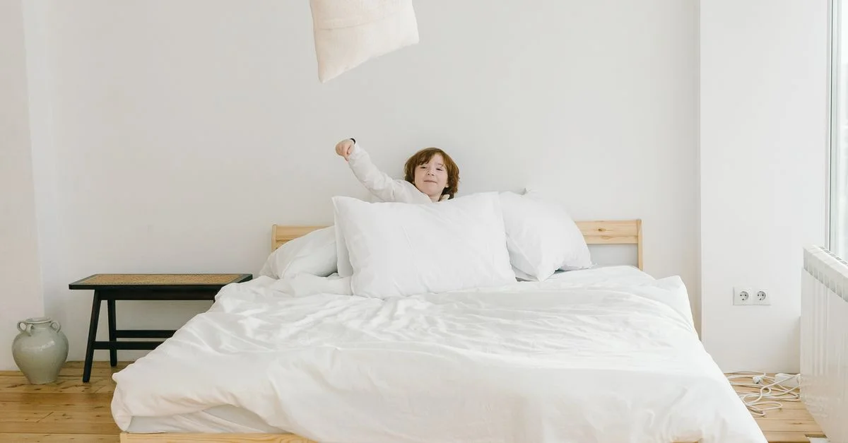 Un jeune garçon lance avec enthousiasme un coussin dans une chambre lumineuse et minimaliste avec une literie blanche.