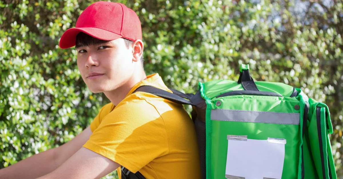 Un livreur asiatique portant une casquette rouge avec un sac isotherme, prêt à livrer en extérieur.