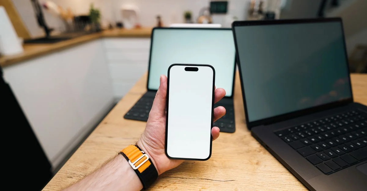 Une main tient un smartphone dans une cuisine contemporaine avec des ordinateurs portables sur une table en bois.